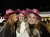 Tre smilende unge kvinner i rosa cowboyhatter foran et opplyst kveldsarrangement.