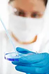 Laboratoriearbeider med munnbind og hansker holder en petriskål og bruker en pipette.