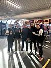 Fem personer poserer i en innendørs racinghall med racingbilder og sikkerhetsbarrierer i bakgrunnen.
