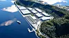 Flyfoto av et stort industrikompleks ved en skogkledd fjord med brygge og båt.
