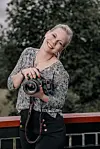 Smilende fotograf med kamera foran trær og et rekkverk.