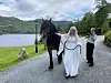 Kvinne i hvit kjole leder en mørk hest på grusvei ved en innsjø og grønne fjell.