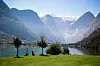 To personer ved en fjord med grønne enger, bratte fjell og snødekte topper.