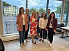 Gruppe voksne samlet ved et stort vindu i et lyst lokale med utsikt mot vannet.