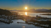 Solnedgang over en kystby med fjord, småbåthavn og fjell.