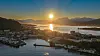 Solnedgang over en kystby med fjord, småbåthavn og fjell.