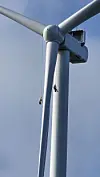 To arbeidere klatrer på en vindturbin under rotorbladene mot blå himmel.