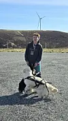Person med to hunder foran en vindturbin i et åpent, kupert landskap.