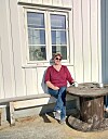Kvinne med solbriller sitter på en benk ved et rustikt bord utenfor en trebygning.