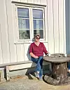 Kvinne med solbriller sitter på en benk ved et rustikt bord utenfor en trebygning.