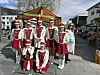 Gruppe i røde og hvite uniformer poserer på et bytorv sammen med en kvinne i turkis kjole.