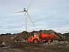 Oransje tippbil i et steinbrudd med en vindturbin i bakgrunnen.