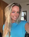 Smilende kvinne med langt blondt hår og blå topp innendørs.