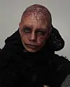Nærbilde av en person med barbert hode, svart antrekk og dramatisk rødlig scenesminke.