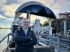 Mann ved en brygge med sjø, båter og en buet overbygning i bakgrunnen.