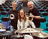Tre musikere med trompeter og trommesett foran seter i en konsertsal.