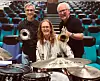 Tre musikere med trompeter og trommesett foran seter i en konsertsal.