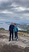 To turgåere står på en fjelltopp med utsikt over tettsted, fjord og fjell.