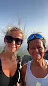 To kvinner med solbriller poserer foran et fjellandskap under blå himmel.