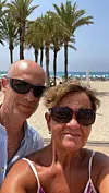 To voksne personer tar en selfie på en solrik strand med palmer og parasoller bak seg.